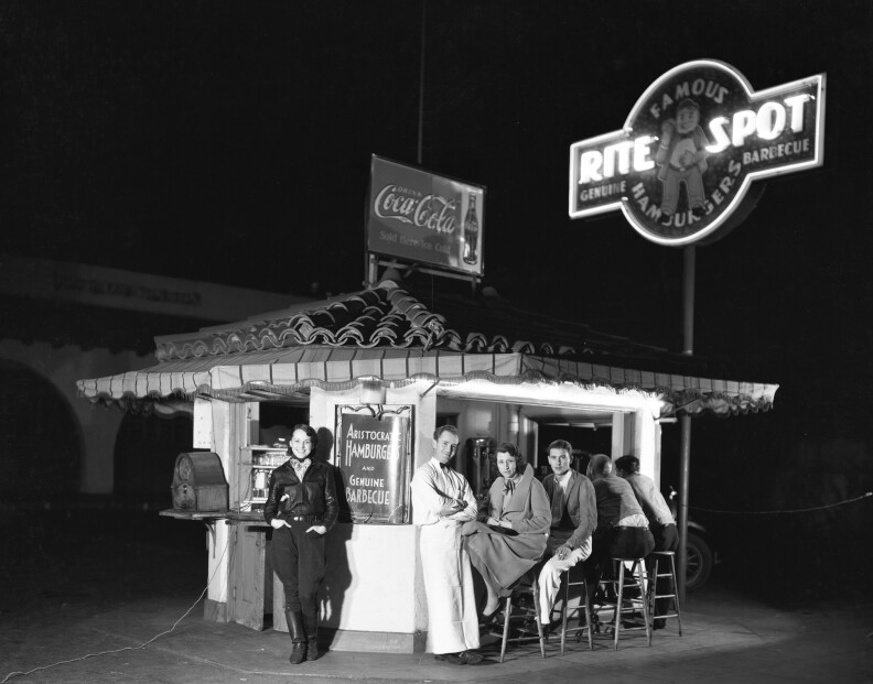 A black and white photo of a road stand from the 1930's. It has a Spanish tile roof, with a sign that says Coca Cola on the top. Next to it is a brightly lit neon sign which says RITE SPOT. Several people are sitting at stools and leaning on the counter, looking at the camera. It's night time.