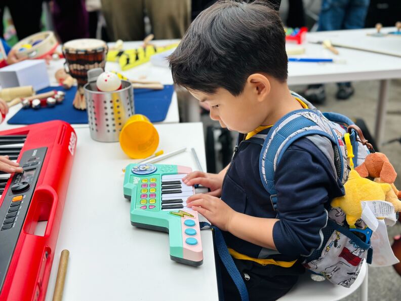 A young boy sits on a small chair and plays a bright green and pink keytar.