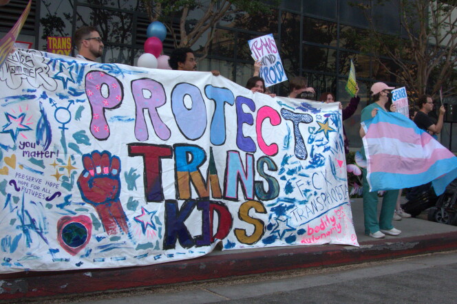Protesters holding a sign that says various slogans in support of trans health care for youth, including, most prominently, "Protect Trans Kids."