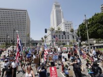 A crowd of people hold signs and American flags as they walk on a street. Buildings are standing behind them with clear skies in the distance.
