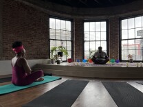 Two brown skinned people sit cross-legged in a dark studio room on yoga mats. 