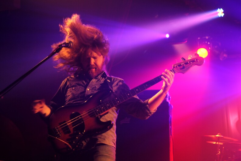 Bassist Reed Mathis flips his long hair on stage while playing guitar. He's under purple and red lighting.