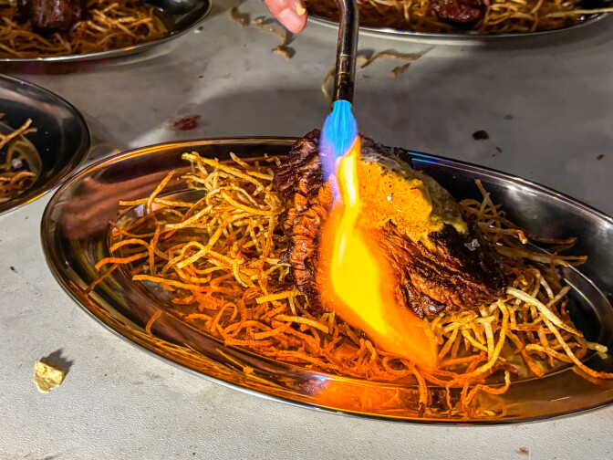 A kitchen torch with a blue and orange flame is held over a hanger steak served on crispy shoestring frites in a silver oval dish, with additional plates visible in the background.