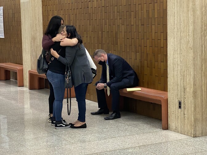 Three people in a group hug in a hallway. On a bench a short distance away a man in a suit and a mask sits with his hands clasped and his shoulders slumped.
