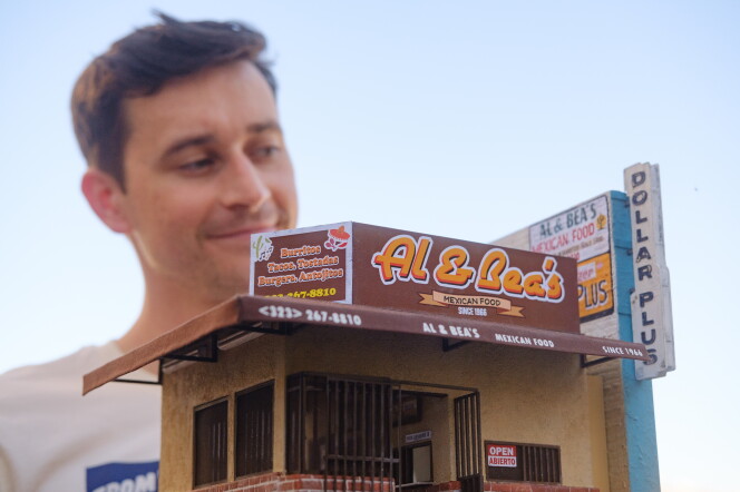 A small version of Al & Bea's Mexican Food in the foreground. It's a brown and tan-colored building with an awning that overlooks a front entrance with metal bars. A small open sign is in the window. On the right side is a wall that has a sign for the business and a Dollar Plus store. The building is being held up by the artist, who is standing in the background.