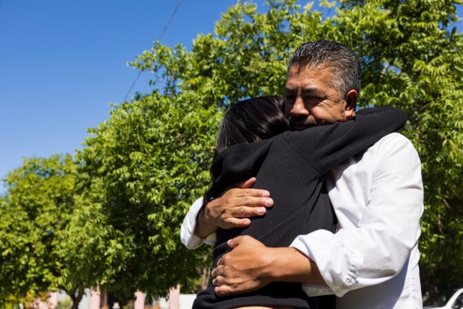 A man wearing a white long sleeved shirt hugs a woman wearing a black, long-sleeved hooded sweater.