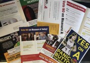 Campaign mailers spread across a table. One has a drawing of a housekeeper and says "Yes on A. Panic Button for Housekeepers." Another says "Vote NO on Measure A Today!"