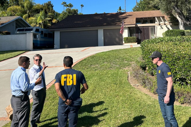 Various men, two with FBI shirts stand at the bottom of a driveway and lawn that leads to a single story house. 