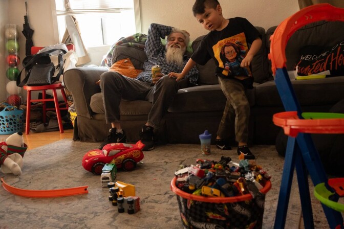 An older man with a large gray beard sits on a couch in a living room, while a small boy plays with toys.