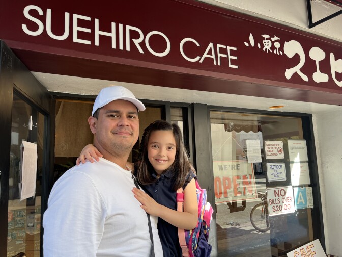 A man in a white cap holding his daughter stands outside a restaurant with an awning that reads "Suehiro Cafe" in English and Japanese.