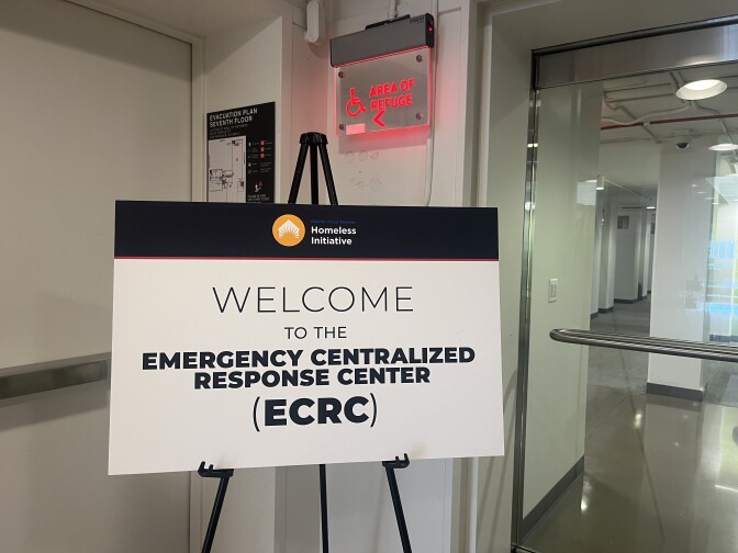 A white sign with a black stripe across the top that says "Homeless Initiative". The sign says "Welcome to the Emergency Centralized Response Center (ECRC). The sign is in the entrance to an office building.