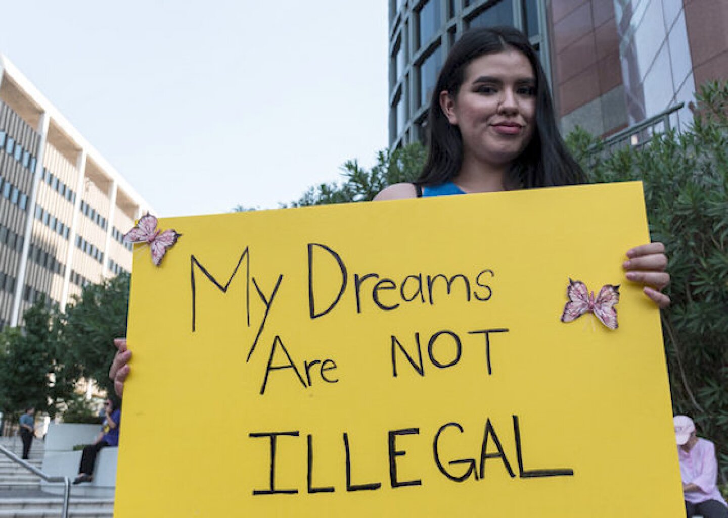 A photo of a woman with black straight hair holding a yellow poster with the words my dreams are not illegal.