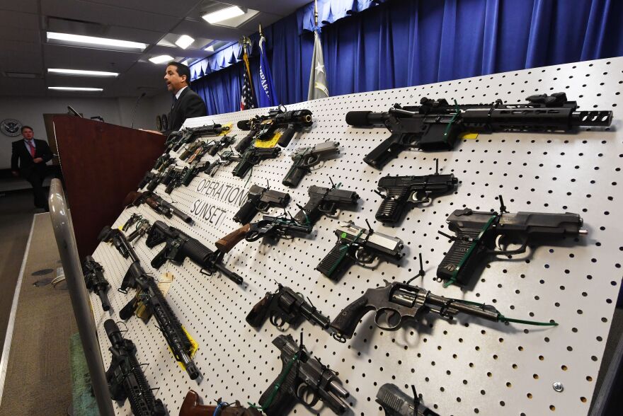 A couple dozen weapons are displayed on a pegboard labeled "Operation Sunset" while a man in a suit speaks at a lectern in the background.