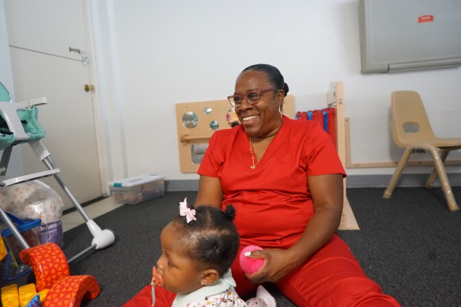 A woman with a dark skin tone and dark hair pulled back into a bun wears pink glasses and red scrubs. She has a big smile and sits on the ground holding a children's toy. A baby with dark skin tone sits in front of her, with a pink bow in her hair.