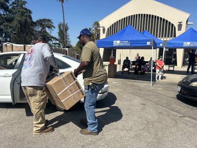 Two middle-aged men wearing baseball caps lift a large box into a white car. In the background are blue shade tents that read "Los Angeles Department of Water and Power." The man on the left has light brown skin and wears a grey long sleeved shirt and khaki work pants and boots and the man on the right is Black and wears a khaki green short sleeved shirt, jeans and work boots. 