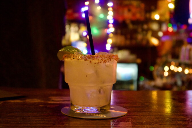 A margarita with a chili-salt rim and lime wedge sits on a bar coaster, with string lights glowing in the background.