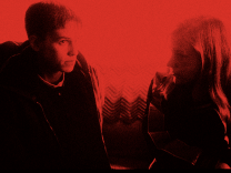 In a scene from the 1999 film “Boys Don’t Cry,” on the left side of the image, actor Hilary Swank plays trans man Brandon Teena. Her brown hair is cut short, and there is a serious expression on her face as she sits on a couch and looks at someone off camera. She is wearing a dark shirt with the collar of a white undershirt showing, and a puffy brown jacket on top. Next to Swank on the couch (covered in a multi-colored, chevron-pattern blanket) actor Chloe Sevigny (playing Brandon’s girlfriend, Lana Tisdel) is seen in profile, looking at Swank. She has long-ish reddish blonde hair and is wearing a black jacket with gray stripes down the arms.