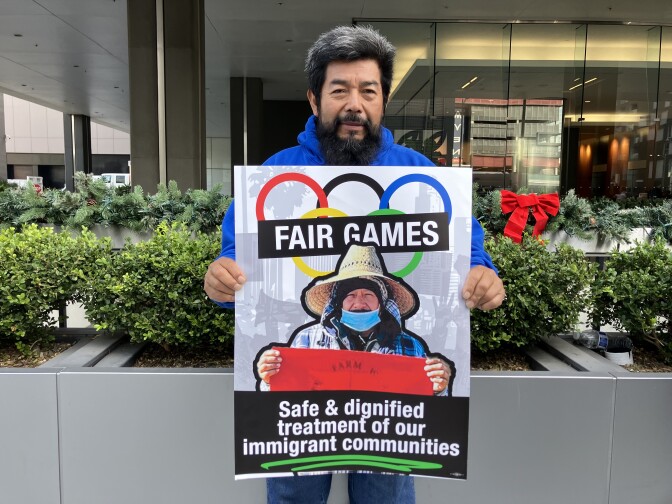 A man with a medium skin tone and black and gray hair wears a blue sweatshirt and holds a poster that reads "FAIR GAMES: Safe and dignified treatment of our immigrant communities."