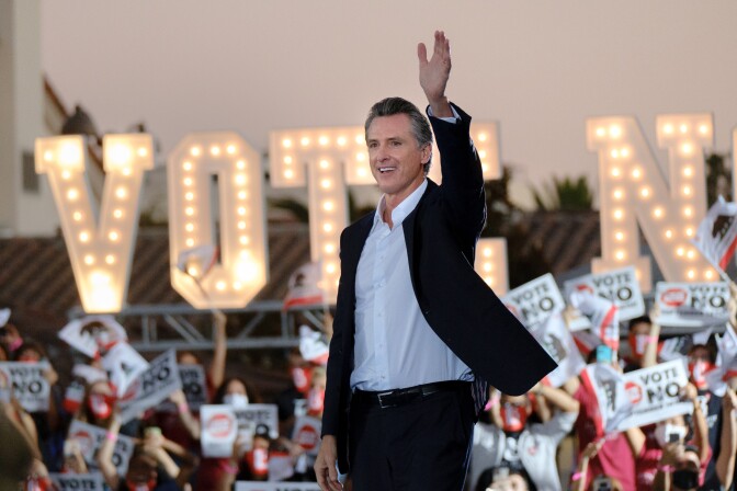 California Governor Gavin Newsom is shown smiling and waving from a stage. Behind him, the word VOTE appears in very large, marquee-style lights. An audience can be seen as well, holding signs that read Vote No. 