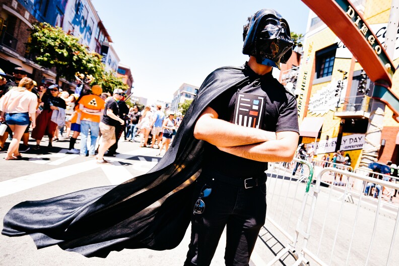 A fan in a Darth Vader costume stands with arms crossed on a San Diego Gaslamp District street, with fans walking in the background, a blue sky, and various businesses decorated for Comic-Con.