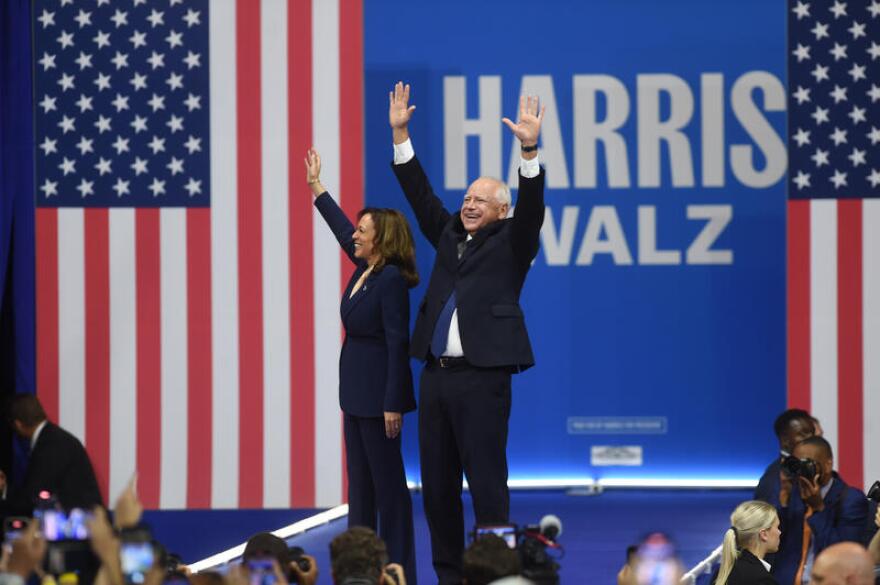 Two people both wearing suits wave to a crowd with an American flag in the background. 