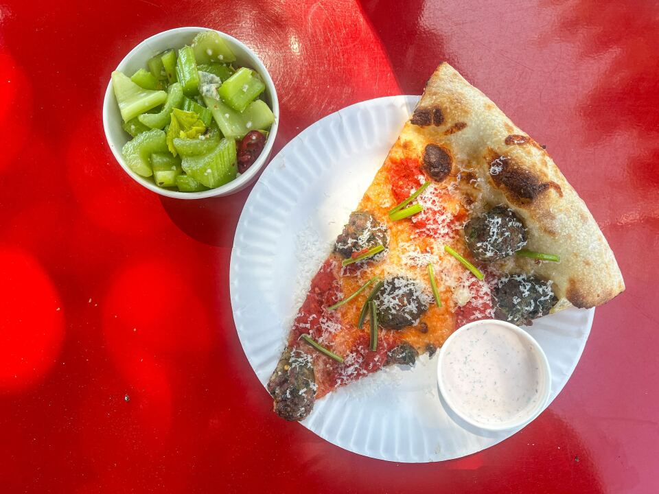 On a white paper plate lies a slice of pizza topped with brown meatballs, green vegetables, and finely shredded white cheese. Adjacent to the pizza is a small container of thick white liquid. At the upper left corner of the plate, there is another small container containing light green sliced celery, with small chunks of blue cheese. The entire scene is set against a red backdrop.