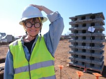 A woman with medium light skin tone, wearing a hard hat and fluorescent vest, stands in front of a stack of gray concrete blocks on a dirt lot in the Los Angeles County neighborhood of Sunset Mesa. 