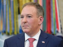 Man with light-toned skin, short brown and gray hair, looking off to the side. He is wearing a dark navy suit jacket over a white collared shirt, a red tie with a small repeating pattern, and a small American flag pin on the lapel.