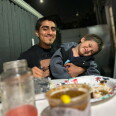 A young man with medium-toned skin smiles as he holds a squirming young boy in his arms at a dinner table. 
