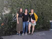 Three light-skinned women stand next to each other in front of a green plant backdrop. The woman to the far left is wearing a brown printed skirt and black top. The woman in the middle is wearing a black top and jeans and the woman to the left is wearing olive shorts with a black top and yellow sweater.