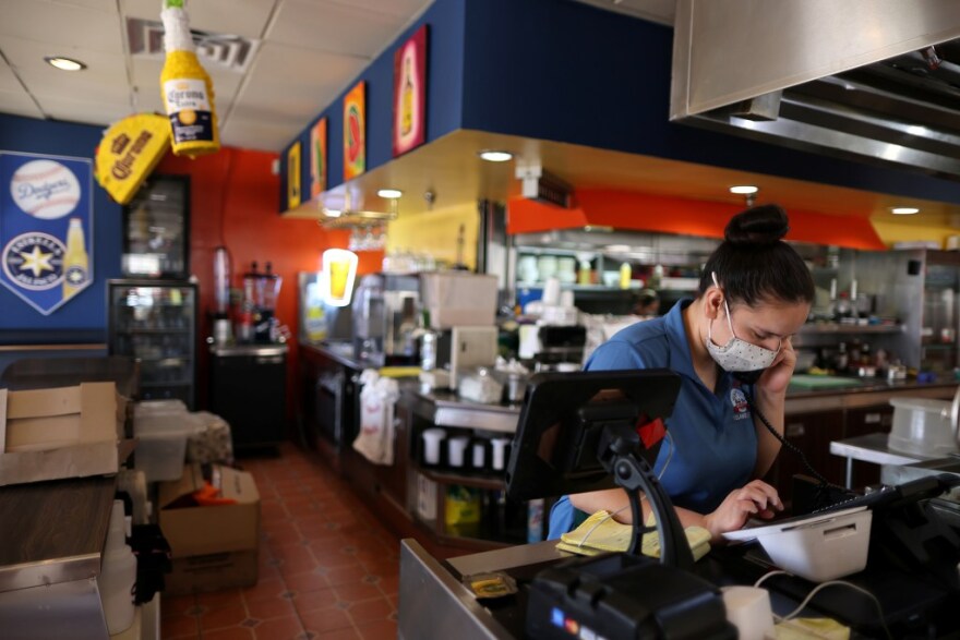 A woman, with her dark hair tied into a bun in a blue collared shirt and white mask, works a cash register at a Mexican restaurant. There is a kitchen in the back.