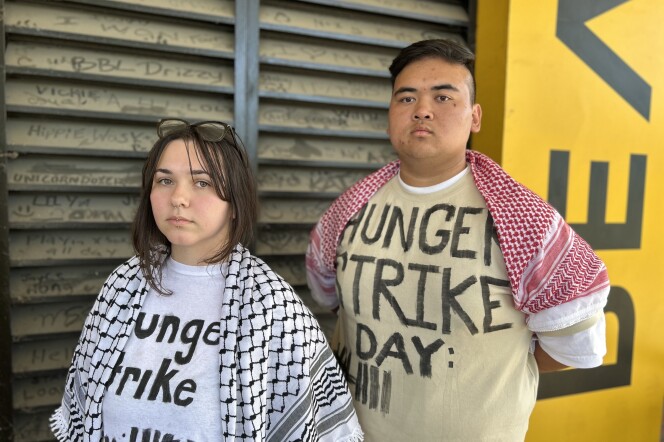 Two students --- one with light skin tone and long dark hair, and one with medium skin tone and short, dark hair --- pose for a photograph while standing next to a campus building. They are clad in keffiyehs,  Under their keffiyehs, their shirts read HUNGER STRIKE DAY 9.