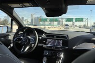The interior of a car taken from the backseat, with notably no driver sitting behind the wheel. The car is driving on a freeway near the downtown Los Angeles skyline in the distance on the left.