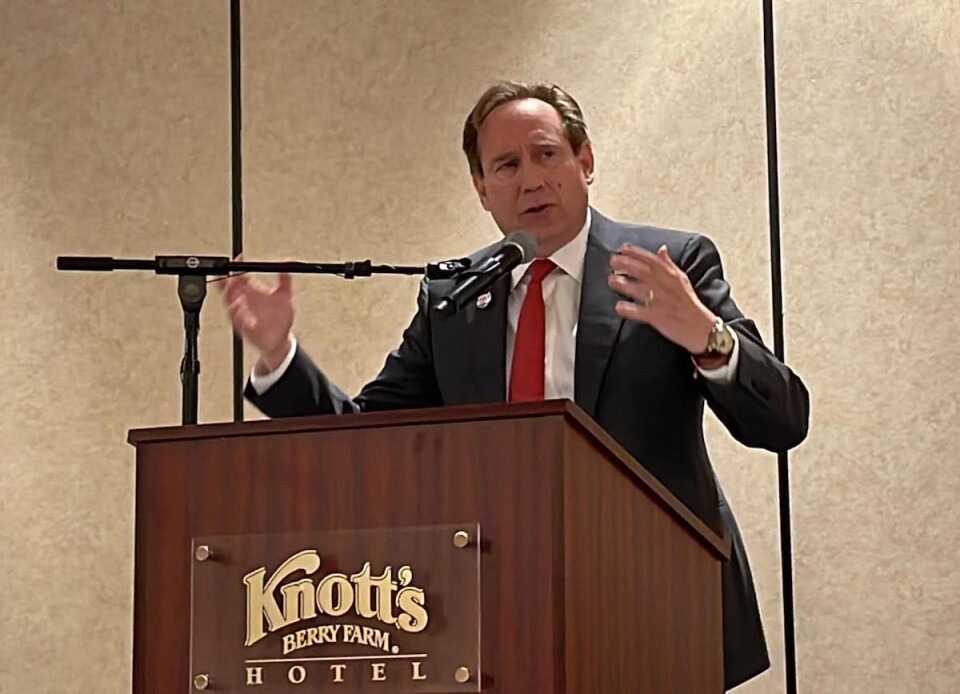 A white man wearing a gray suit and red tie speaks into a microphone in front of a brown podium bearing a sign that reads "Knott's Berry Farm Hotel."