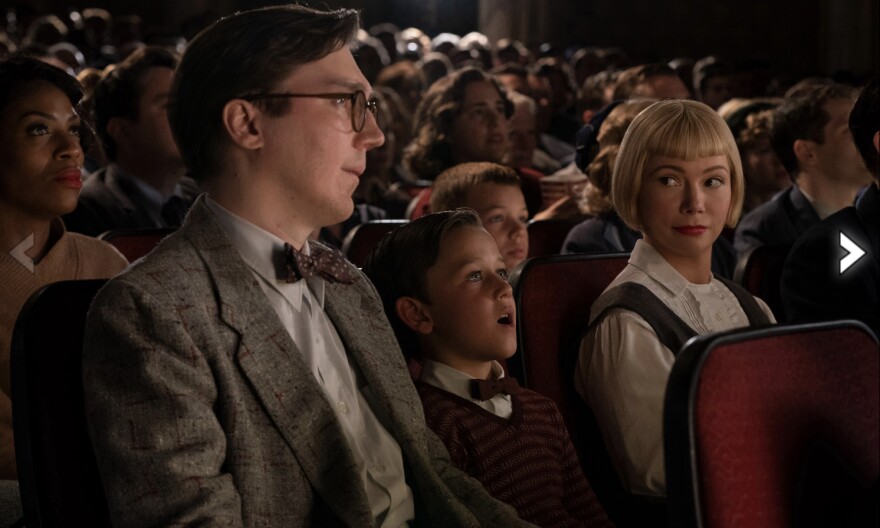 A light-skinned man with glasses. 1950's jacket and bowtie sits in a packed movie theater next to a young light-skinned boy wearing a sweater and a bowtie. Next to him is a light-skinned woman with a blond bob and bangs. She's looking at the man. The young boy is looking entranced at the screen.