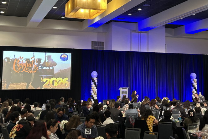 A hotel ballroom filled with dozens of dinner tables and a stage lined with bright blue curtains. A screen is set up on the left of the stage with an image that reads "Congrats Class of 2026." A pair of white, black and gold balloon pillars are set up on either side of a silver podium toward the center of the stage.