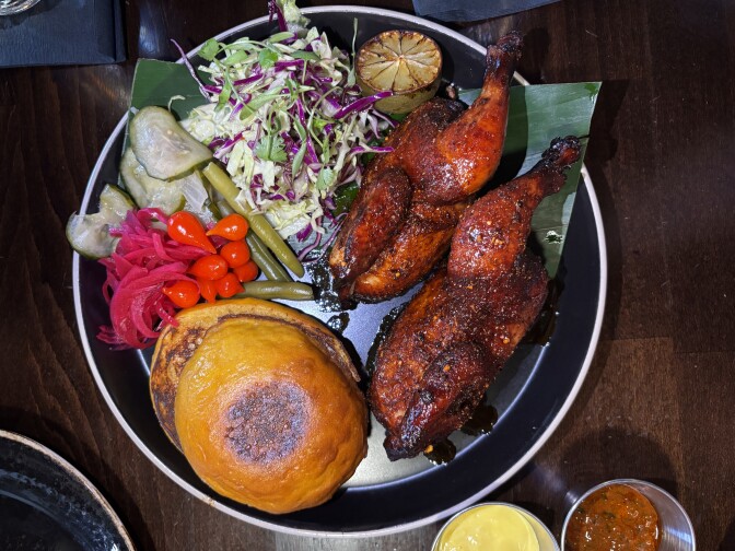A full plate built for tearing and sharing — smoky roast chicken, tangy pickles, and a golden round of a sweet potato bun. 