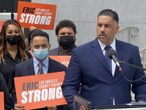 LA Sheriff candidate Eric Strong stands at a microphone wearing a blue suit with a white shirt and blue tie with tiny white dots. Three people stand to the left; on the far left is a woman with long auburn-red hair wearing a black face mask, in the middle is a man wearing a blue suit, light blue shirt and grey tie, wearing a light blue face mask, holding an orange sign that says Eric Strong, Los Angeles County Sheriff. The word "Eric" is in black and the rest of the words are in white. Above his head, someone's hand is holding the same sign. On the right is a man wearing a dark suit and shirt who is also holding an Eric Strong sign, but only the "Los A" portion is visible. 