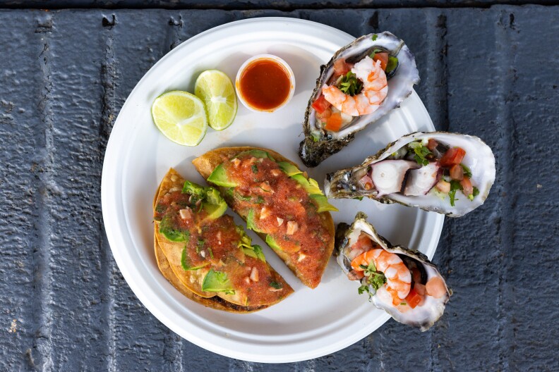 A white paper plate sits atop a gray concrete surface. On the plate are two crispy tacos that are accented with slices of avocado and sathered in a red sauce containing bits of cooked pink shrimp. The tacos sit next to three oysters that have been topped off with pieces of sliced octopus and pink, cooked shrimp tails. Next to the tacos and oysters, there are two slices of limes and a cup of red salsa.