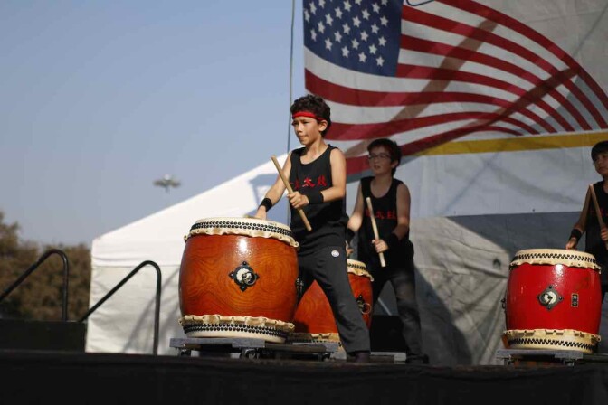 Two boys play taiko drums with an American flag waving behind them.