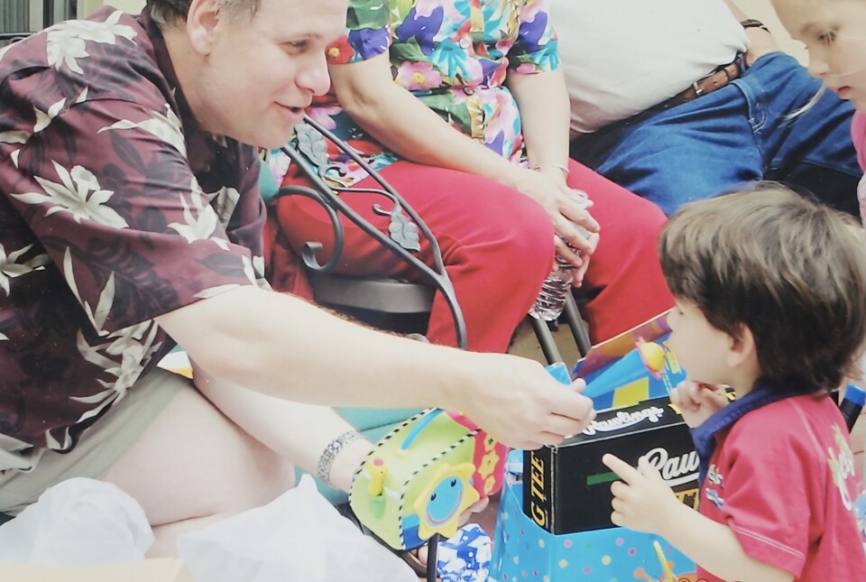 A side profile shot of fair skinned man in a dark reddish shirt with flowers holds a plastic toy microphone in front of a young dark haired, fair skinned chid