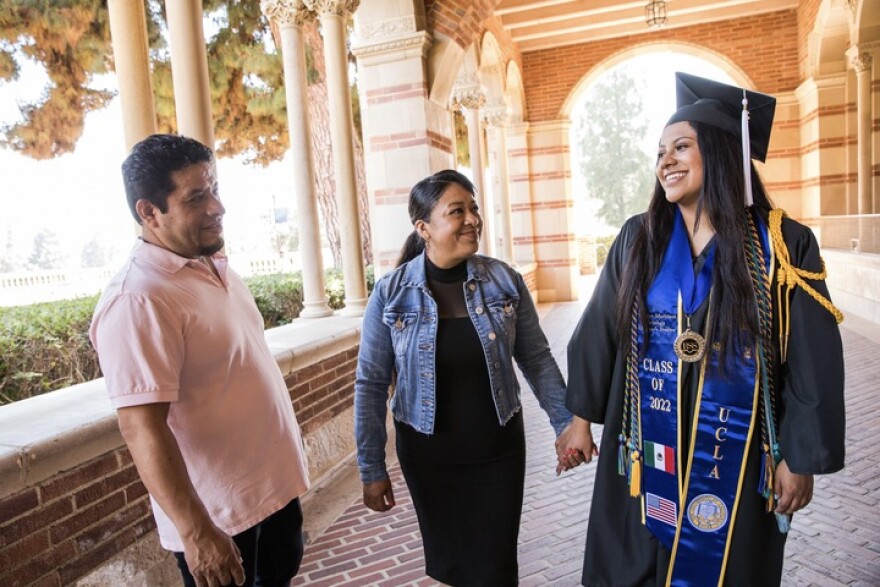 On the right, a woman with medium skin tone and long dark hair wears a black cap and gown. Her blue stole says "UCLA" and "Class of 2022." It also features a Mexican flag and a flag of the U.S. She holds the hand of a woman with medium skin tone who has her hair in a ponytail. On the left, a man with medium skin tone, clad in a polo shirt, smiles in their direction. The women are also smiling. 