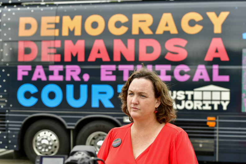 Rep. Katie Porter, a woman with light skin tone wearing an vermilion-colored shirt with a button, stands in front of a black bus with text on it that reads "Democracy demands a fair, ethical court."