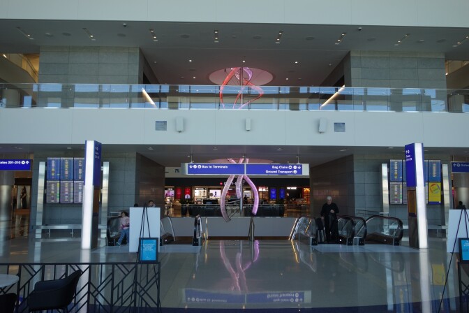 An airport terminal with escalators and a second-story walkway visible. A light-up pink and red sculpture is in the center of the picture.