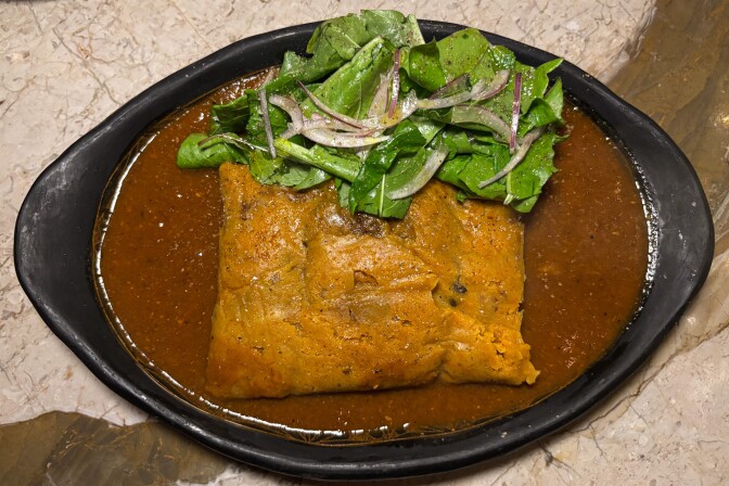 A sauced tamal served in a shallow pool of rich red chile sauce, topped with fresh greens and sliced red onion.