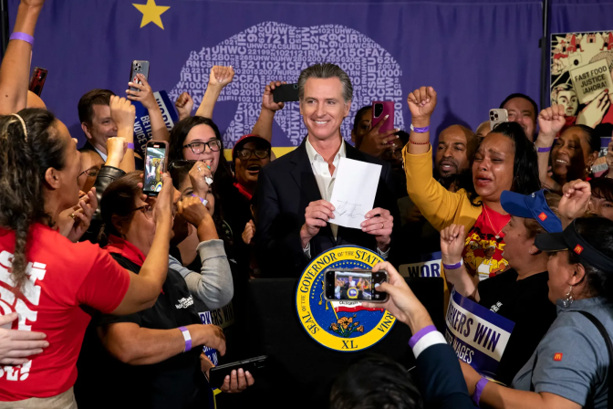 A large crowd of people, many with their fists raised, gather around Governor Gavin Newsom, who is holding a piece of paper and standing in front of a podium with a round logo that reads Seal of the Governor of the State of California
