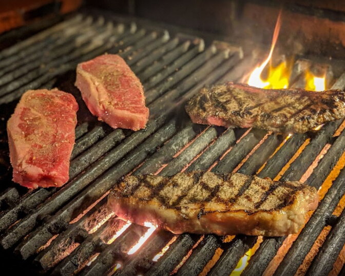 Four raw and grilled steaks cooking on a flaming charcoal grill, two seared with grill marks and two still raw.