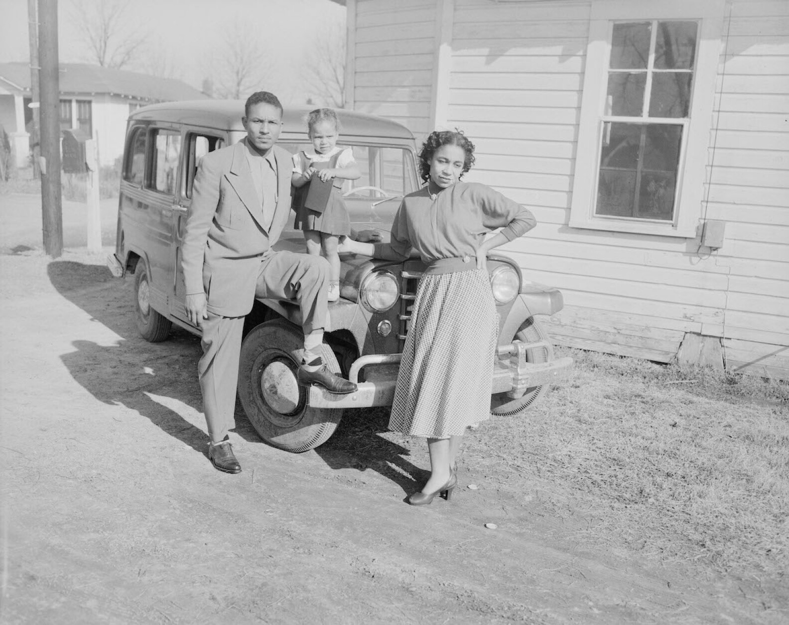 A black and white image shows two adults and a young girl leaning against a mid-20th century car.