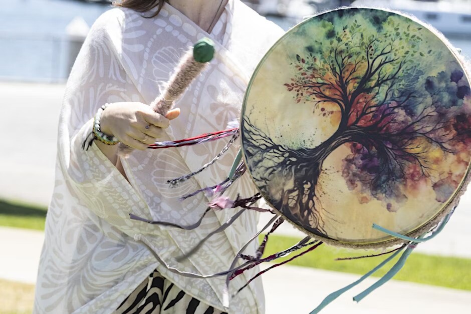 A person wearing a white patterned shawl holds a decorated drum and mallet. The drum features an artistic depiction of a tree with colorful leaves, and several ribbons hang from the drum's edge.