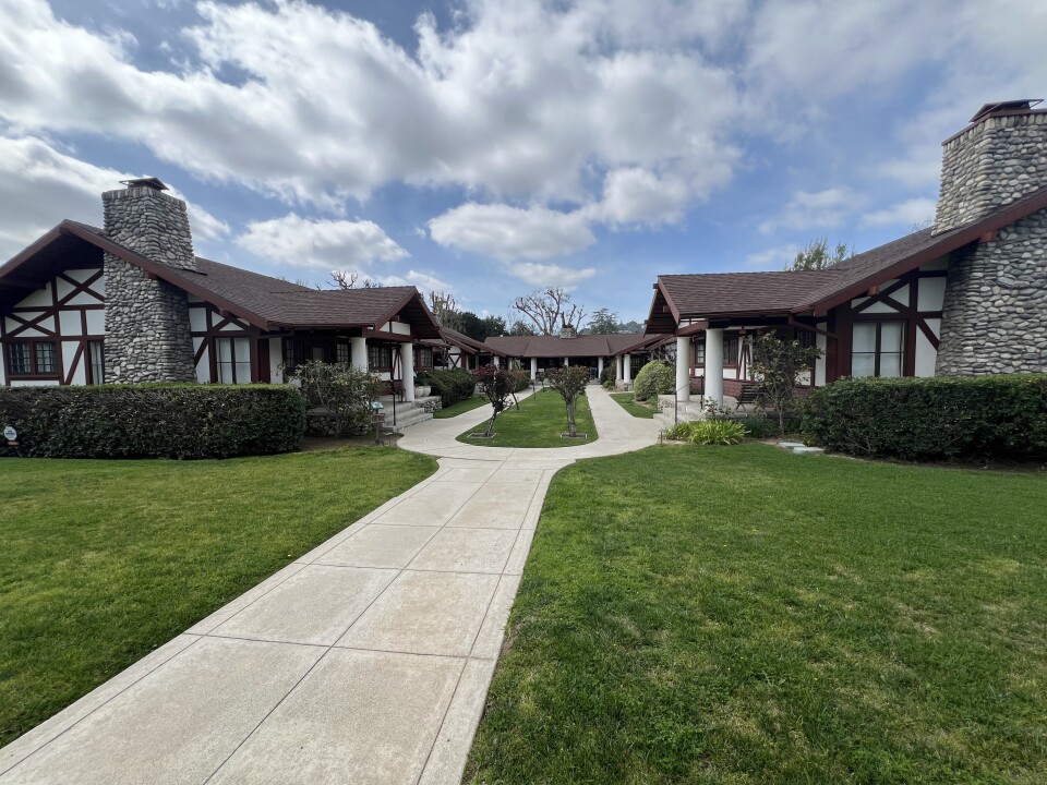 A single cement walkway on a lawn splits into two in the center courtyard section of a multi-family housing complex. It's made up of six identical single story homes. They have brown sloped roofs, white outer walls with decorative dark brown criss-crossed beams, two white columns flanking the porch steps, and chimneys covered in river rocks in various shades of grey.